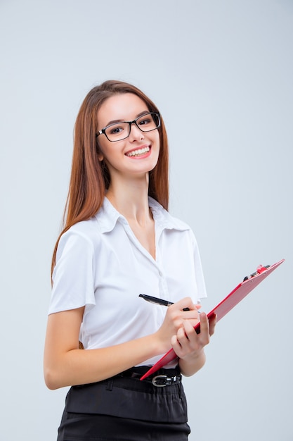 Uśmiechnięta młoda biznesowa kobieta w okularach z piórem i tabletem na notatki na szaro