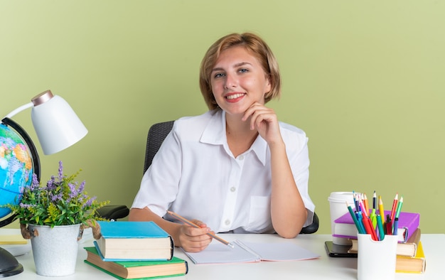 Uśmiechnięta młoda blond studentka siedząca przy biurku ze szkolnymi narzędziami trzymająca ołówek dotykający podbródka, patrząca na kamerę odizolowaną na oliwkowozielonej ścianie