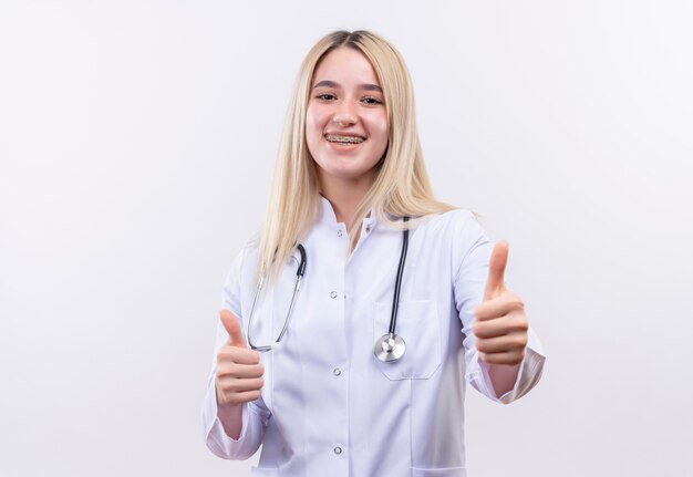 Uśmiechnięta młoda blondynka lekarz ubrana w stetoskop i fartuch medyczny w ortezie dentystycznej kciuki do góry na na białym tle