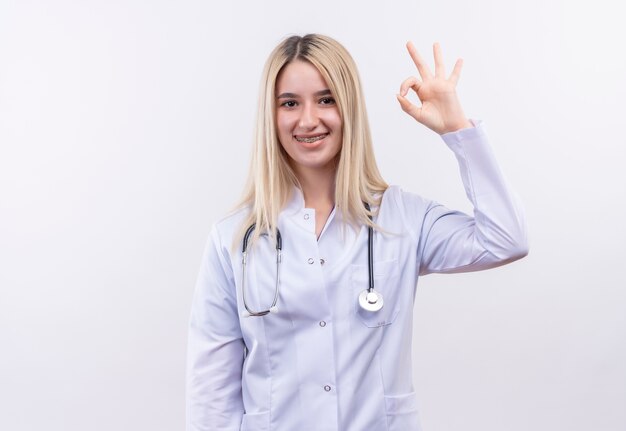 Uśmiechnięta młoda blondynka lekarz ubrana w stetoskop i fartuch medyczny w ortezie dentystycznej pokazujący dobry gest na na białym tle