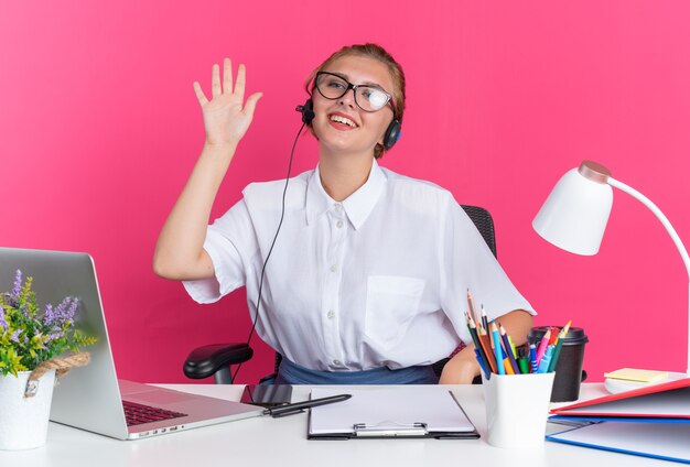 Bezpłatne zdjęcie uśmiechnięta młoda blondynka z call center w słuchawkach i okularach, siedząca przy biurku z narzędziami do pracy, patrząca na bok, wykonująca gest cześć na białym tle na różowej ścianie