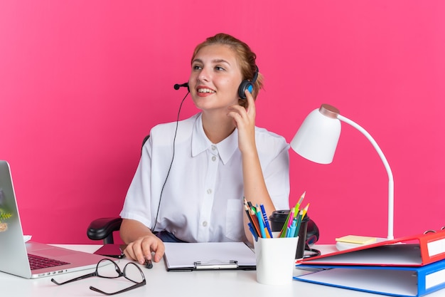 Uśmiechnięta młoda blondynka z call center w zestawie słuchawkowym siedząca przy biurku z narzędziami do pracy dotykającymi zestawu słuchawkowego patrząc na bok at