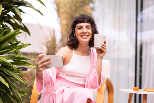 Uśmiechnięta młoda brunetka kaukaska patrząca na kamerę trzymającą telefon i filiżankę siedzącą na zewnątrz Koncepcja stylu życia technologia szczerych emocji