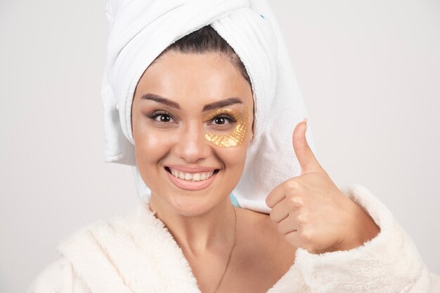 Uśmiechnięta młoda brunetka kobieta ubrana w szlafrok z kosmetycznymi opaskami na oczy.