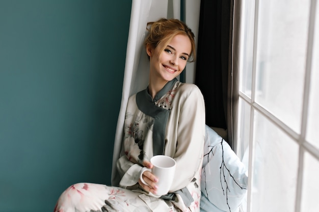 Uśmiechnięta młoda dama z filiżanką kawy, herbatą w niej miała, siedząc na parapecie, dzień dobry relaks. Ubrana w jedwabną piżamę w kwiaty, ma blond włosy. Zdjęcie w turkusowych kolorach.