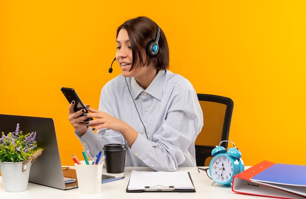 Uśmiechnięta młoda dziewczyna call center na sobie zestaw słuchawkowy siedzi przy biurku i patrząc na telefon komórkowy samodzielnie na pomarańczowej ścianie