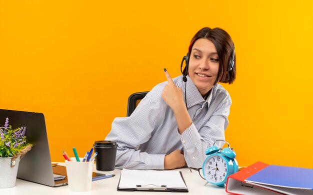 Uśmiechnięta młoda dziewczyna call center noszenie zestawu słuchawkowego siedzi przy biurku patrząc na bok, wskazując za na białym tle na pomarańczowej ścianie