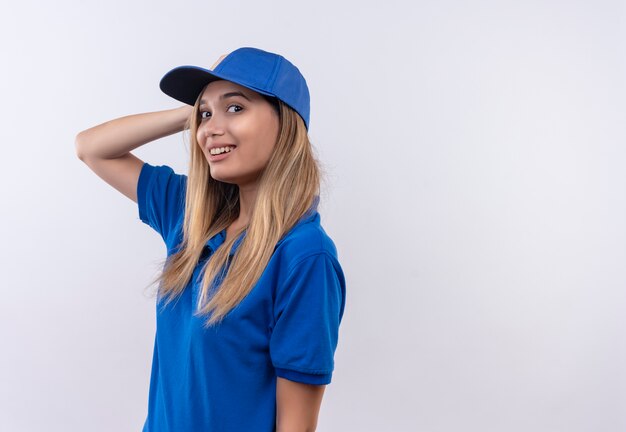 Uśmiechnięta młoda dziewczyna dostawy na sobie niebieski mundur i czapkę kładąc rękę na głowie na białym tle