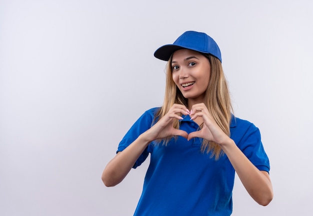 Uśmiechnięta młoda dziewczyna dostawy ubrana w niebieski mundur i czapkę pokazującą gest serca na białym tle na białej ścianie z miejsca na kopię