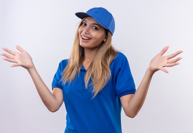 Uśmiechnięta młoda dziewczyna dostawy ubrana w niebieski mundur i czapkę rozkłada ręce na białym tle na białej ścianie z miejsca na kopię