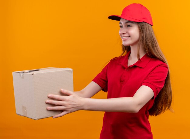 Uśmiechnięta młoda dziewczyna dostawy w czerwonym mundurze, wyciągając pudełko po lewej stronie na odizolowanej pomarańczowej ścianie
