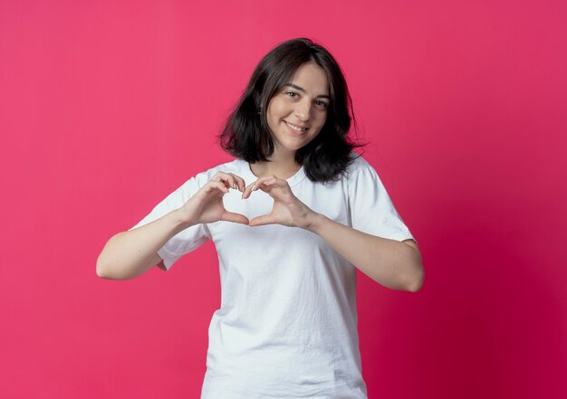 Uśmiechnięta młoda dziewczyna kaukaski robi znak serca w aparacie na białym tle na szkarłatnym tle z miejsca na kopię