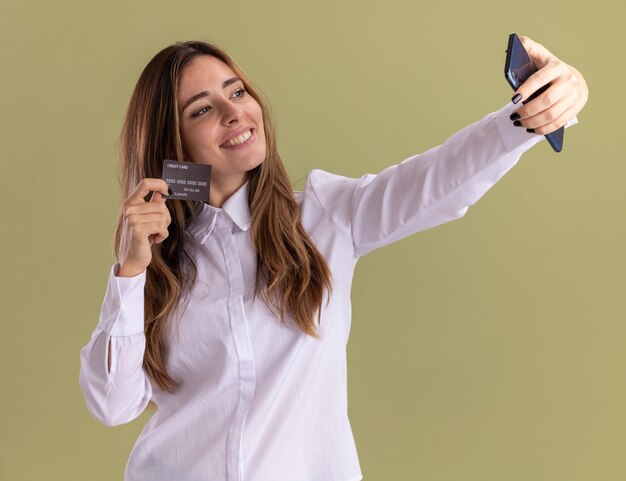 Uśmiechnięta młoda dziewczyna kaukaski trzyma kartę kredytową, trzymając i patrząc na telefon, biorąc selfie na oliwkowej zieleni