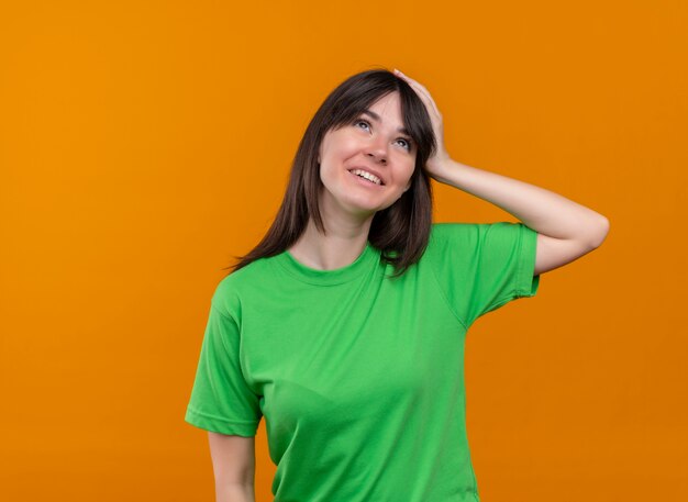 Uśmiechnięta młoda dziewczyna kaukaski w zielonej koszuli kładzie rękę na głowie i patrzy na pojedyncze pomarańczowe tło z miejsca na kopię