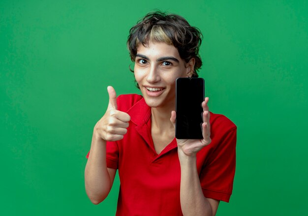 Uśmiechnięta młoda dziewczyna kaukaski z fryzurą pixie trzymając telefon komórkowy i pokazując kciuk w górę na białym tle na zielonym tle z miejsca na kopię