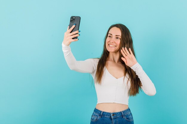 Uśmiechnięta młoda dziewczyna robi selfie, pokazując gest cześć na niebieskim tle