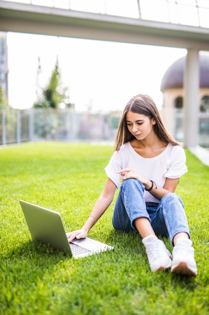 Uśmiechnięta młoda dziewczyna siedzi na trawie z laptopem