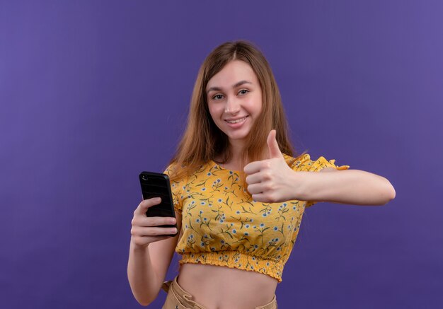 Uśmiechnięta młoda dziewczyna trzyma telefon komórkowy i pokazuje kciuk do góry na odosobnionej fioletowej ścianie z miejsca na kopię
