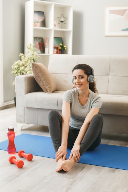 Uśmiechnięta młoda dziewczyna w słuchawkach ćwiczących na macie do jogi przed sofą w salonie