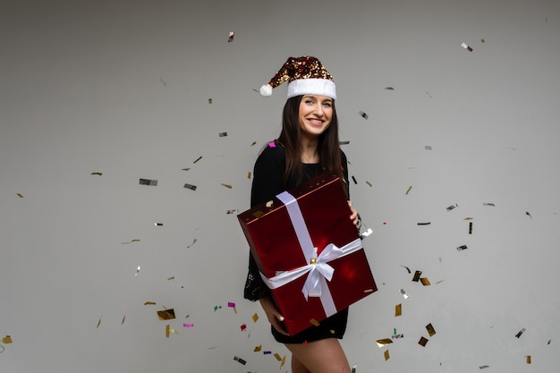 Uśmiechnięta młoda dziewczyna z dużym świątecznym prezentem wskazująca ręką na pustej przestrzeni z świątecznym konfetti na szarym tle