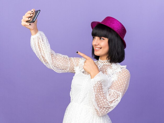 Uśmiechnięta młoda imprezowa dziewczyna w imprezowym kapeluszu stojąca w widoku profilu, biorąca selfie wskazująca na telefon odizolowany na fioletowej ścianie