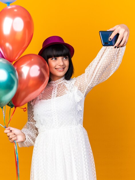 Uśmiechnięta młoda imprezowa kobieta w imprezowym kapeluszu, trzymająca balony, biorąca selfie na pomarańczowej ścianie