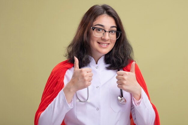 Uśmiechnięta młoda kaukaska dziewczyna superbohatera w czerwonej pelerynie, ubrana w mundur lekarza i stetoskop w okularach pokazujących kciuki do góry na białym tle na oliwkowozielonej ścianie