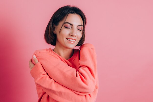 Uśmiechnięta młoda kobieta 20s w ubranie na białym tle na różowym tle portret studio. Koncepcja miłości. Trzymając oczy zamknięte, trzymając ręce skrzyżowane, obejmując się