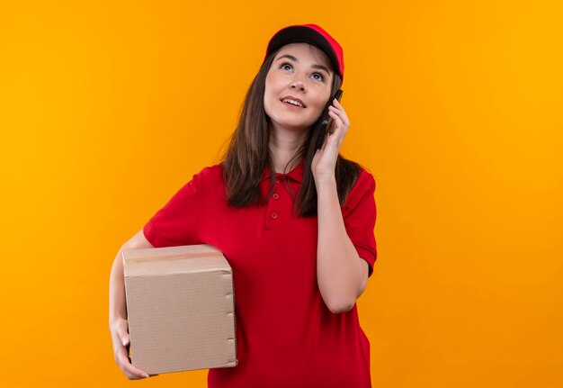 Uśmiechnięta młoda kobieta dostawy na sobie czerwoną koszulkę w czerwonej czapce trzyma pudełko i nawiązywanie połączenia telefonicznego na odizolowanych pomarańczowej ścianie