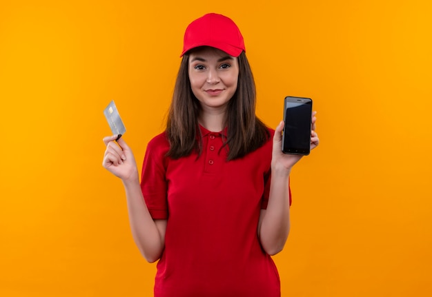 Uśmiechnięta młoda kobieta dostawy na sobie czerwoną koszulkę w czerwonej czapce, trzymając kartę i telefon na odizolowanej pomarańczowej ścianie