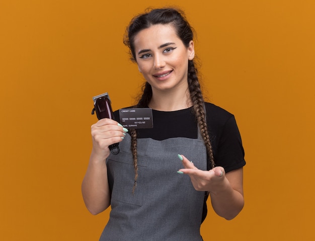 Uśmiechnięta młoda kobieta fryzjer w mundurze gospodarstwa i wskazuje na kartę kredytową i maszynki do strzyżenia włosów na białym tle na pomarańczowej ścianie