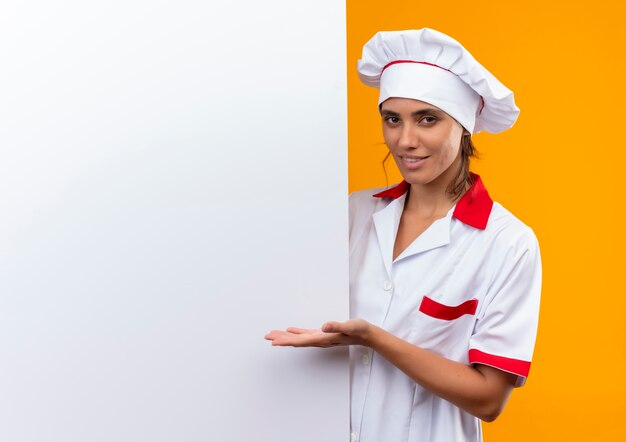 Uśmiechnięta młoda kobieta kucharz ubrana w mundur szefa kuchni trzymając i pokazując ręką białą ścianę z miejsca na kopię