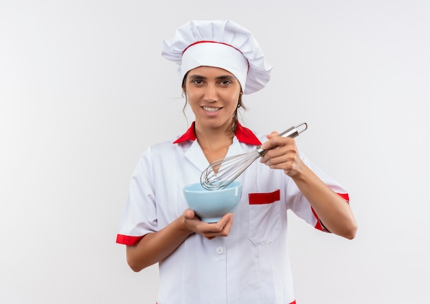 Uśmiechnięta młoda kobieta kucharz ubrana w mundur szefa kuchni trzymając trzepaczkę i miskę z miejsca na kopię