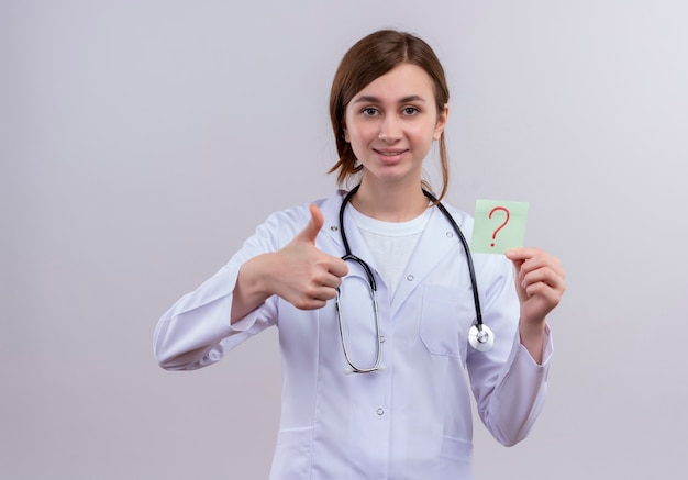 Uśmiechnięta młoda kobieta lekarz ubrana w medyczny szlafrok i stetoskop pokazując kciuk do góry i trzymając papierową notatkę ze znakiem zapytania na odosobnionej białej ścianie