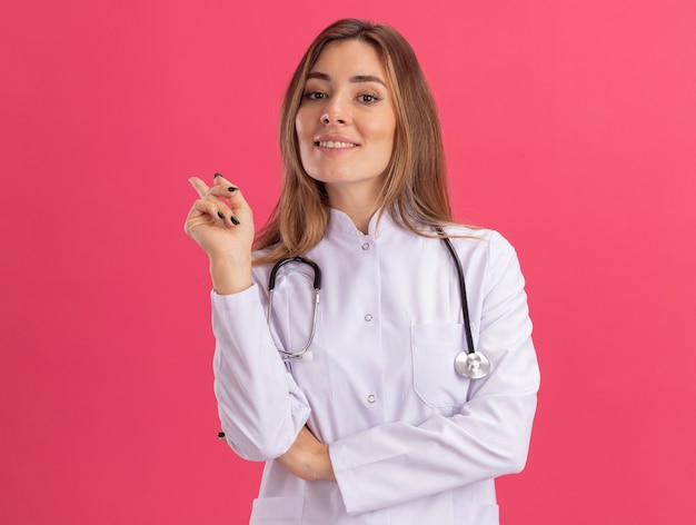 Uśmiechnięta młoda kobieta lekarz ubrana w szatę medyczną z punktami stetoskopu po stronie na białym tle na różowej ścianie z miejsca na kopię