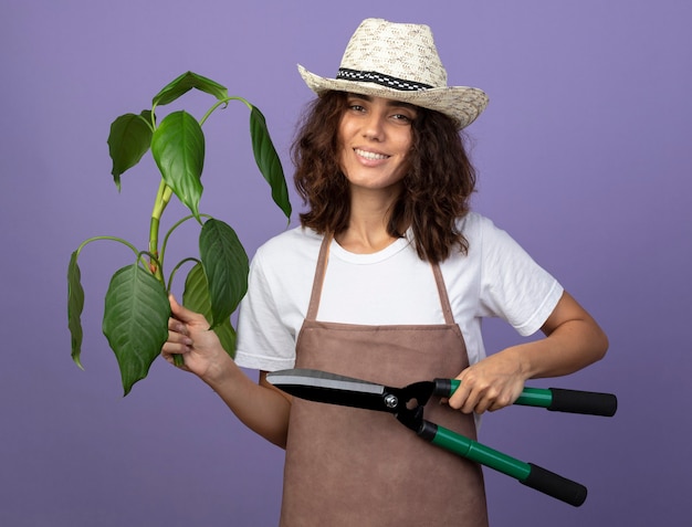 Bezpłatne zdjęcie uśmiechnięta młoda kobieta ogrodnik w mundurze na sobie kapelusz ogrodniczy gospodarstwa roślin z maszynkami do strzyżenia