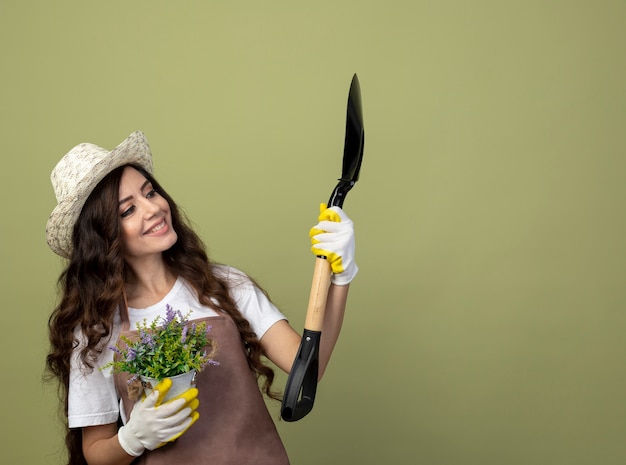 Uśmiechnięta młoda kobieta ogrodnik w mundurze na sobie kapelusz ogrodniczy i rękawiczki trzyma doniczkę i patrzy na łopatę na białym tle na oliwkowej ścianie