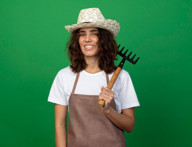 Uśmiechnięta młoda kobieta ogrodnik w mundurze na sobie kapelusz ogrodniczy stawiając prowizję na ramieniu