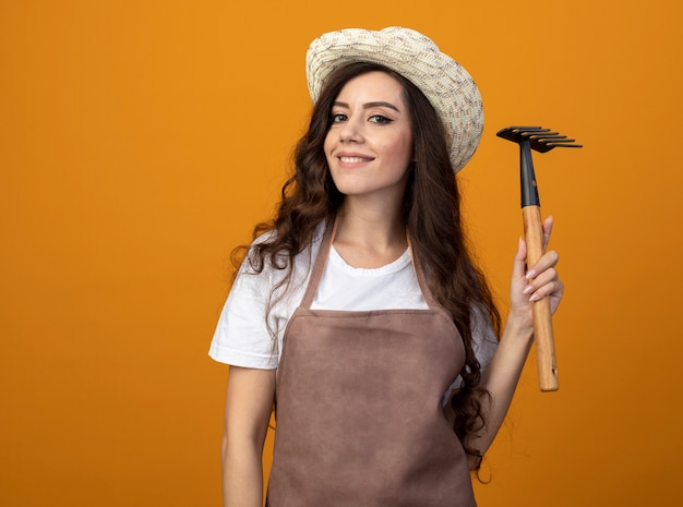 Uśmiechnięta młoda kobieta ogrodnik w mundurze na sobie kapelusz ogrodniczy trzyma prowizję na białym tle na pomarańczowej ścianie z miejsca na kopię