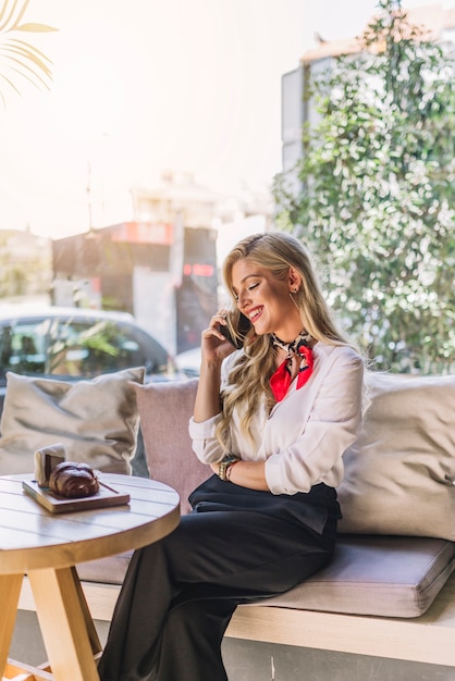 Uśmiechnięta młoda kobieta opowiada na telefonie komórkowym podczas gdy siedzący w caf�