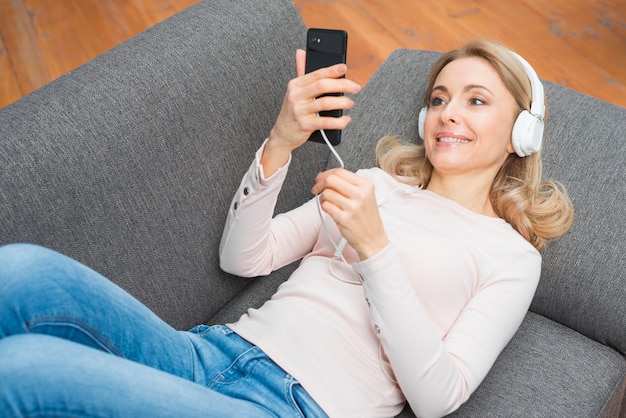 Uśmiechnięta młoda kobieta patrzeje telefon komórkowy słuchającą muzykę na hełmofonie