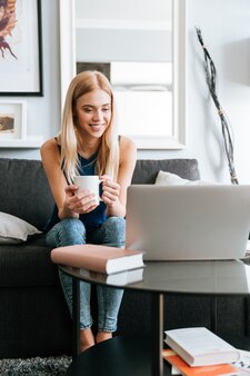 Uśmiechnięta młoda kobieta pije kawę i używa laptop na kanapie