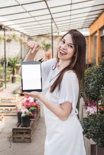 Uśmiechnięta młoda kobieta pokazuje cyfrową pastylkę z pustym bielu ekranem w szklarni