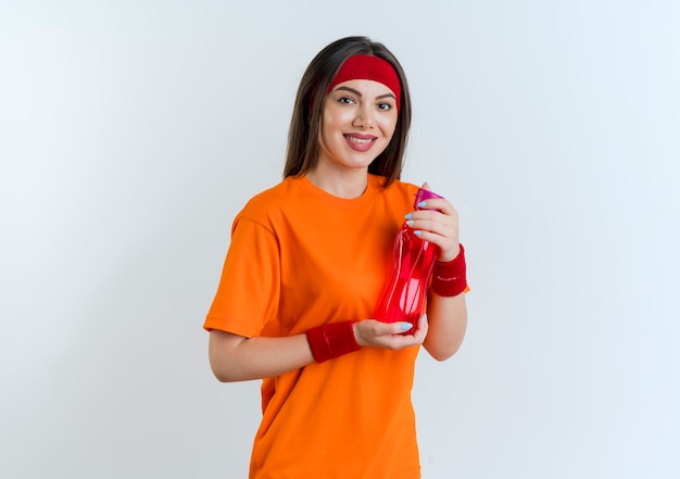 Uśmiechnięta młoda kobieta sportowy noszenie opaski i opaski na rękę trzymając butelkę wody patrząc na białym tle
