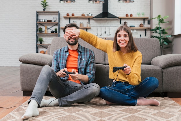 Uśmiechnięta młoda kobieta trzyma joystick zakrywający jej męża oczy podczas gdy bawić się wideo grę