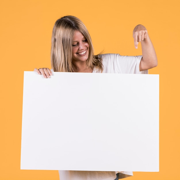 Uśmiechnięta młoda kobieta wskazuje palec wskazującego przy białym pustym plakatem