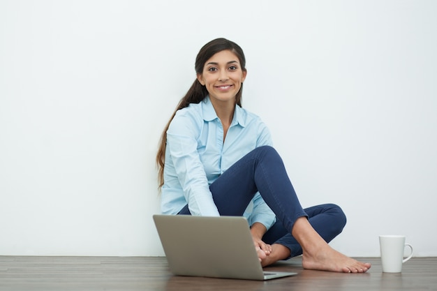 Uśmiechnięta młoda kobieta z laptopem na podłodze i herbata