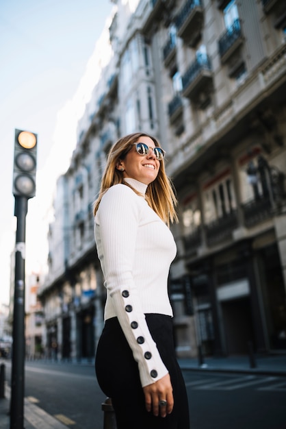 Bezpłatne zdjęcie uśmiechnięta młoda kobieta z okularami przeciwsłonecznymi blisko drogi w mieście