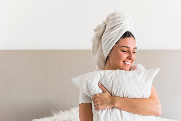 Uśmiechnięta młoda kobieta zawija jej głowę z ręcznikiem trzyma białą poduszkę