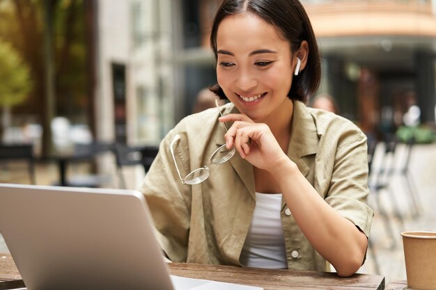 Uśmiechnięta młoda koreańska kobieta patrzy na ekran swojego laptopa z zadowoloną miną, pracuje zdalnie z zewnątrz
